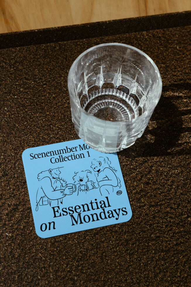 Glass tumbler on textured surface next to blue coaster featuring cartoon characters and text Essential Mondays, ideal for mockup and graphic design projects.