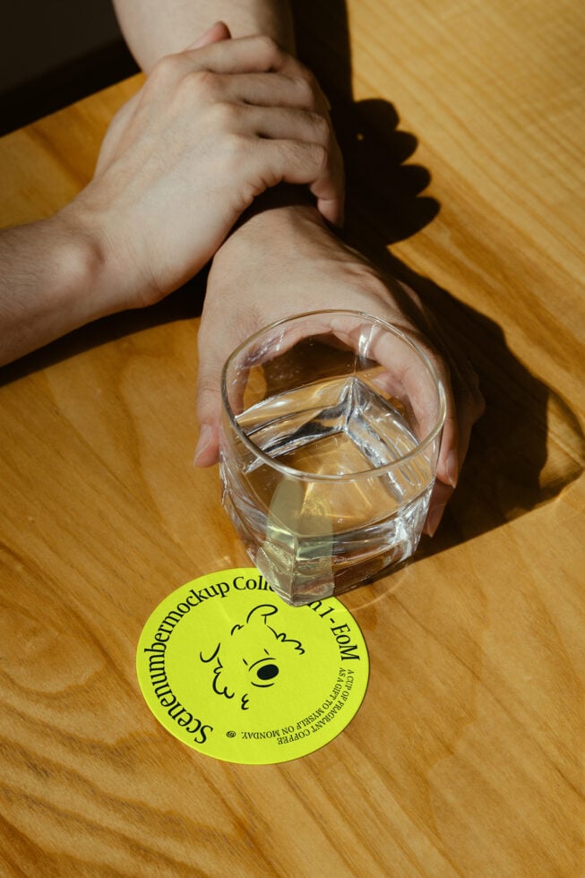 Hand holding a glass above a yellow round coaster mockup on a wooden table perfect for designers creating drinkware branding templates.