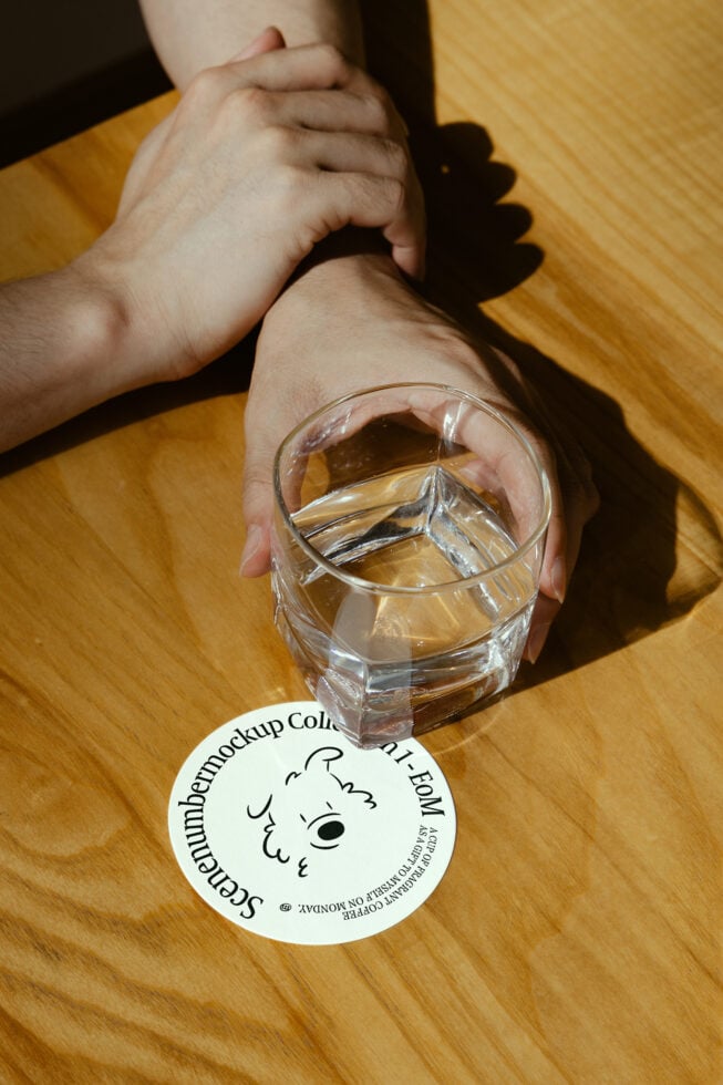 Hands holding a glass of water on a wooden table with a round paper coaster mockup featuring a doodle design. Perfect for branding and graphic designers.