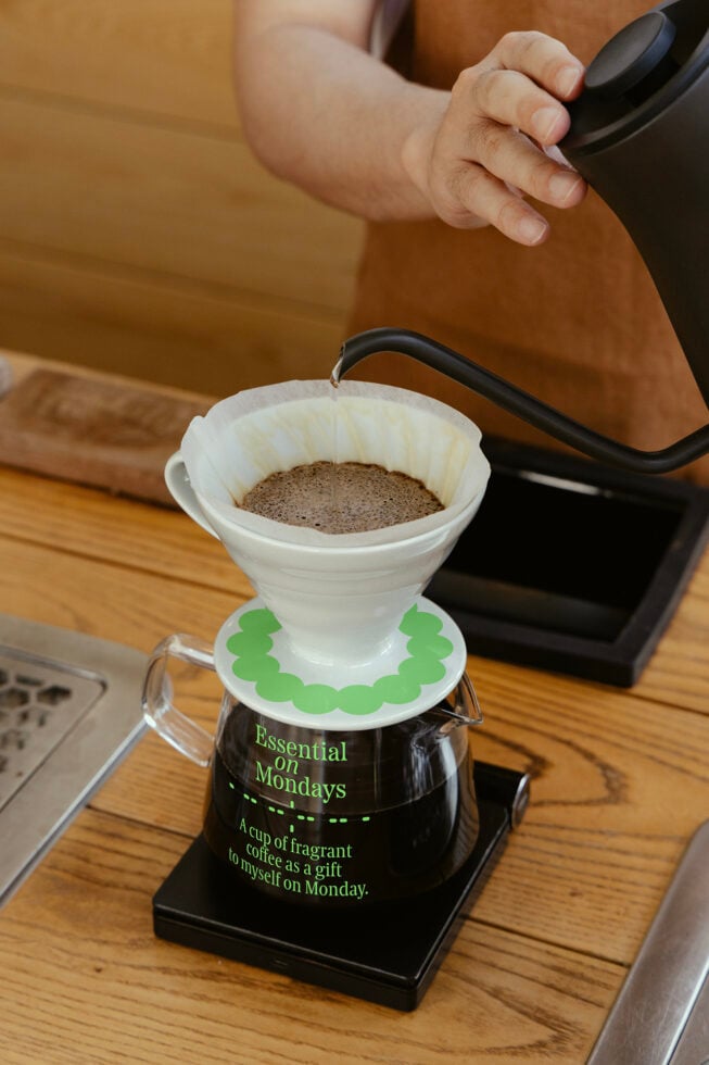 Person brews coffee using pour-over method in a modern kitchen setting. Focus on drip filter with text Essential on Mondays. Keywords: coffee, brewing, kitchen.