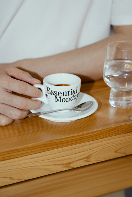 Hand holding white espresso cup with Essential on Monday text, saucer with spoon