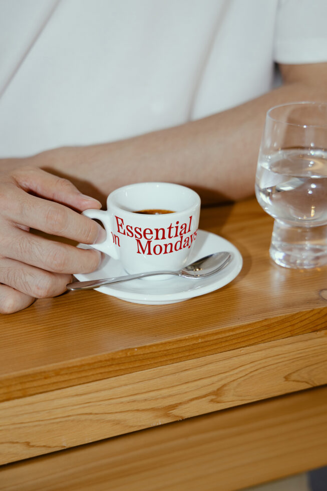 Hand holding espresso cup on wooden table with Essential Mondays text, next to water glass. Perfect for mockup, branding, and design projects.