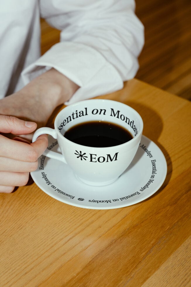 Coffee cup mockup with EoM and Essential on Mondays text. Hand holding cup over a saucer on a wooden table. Ideal for designers, branding, product mockup.