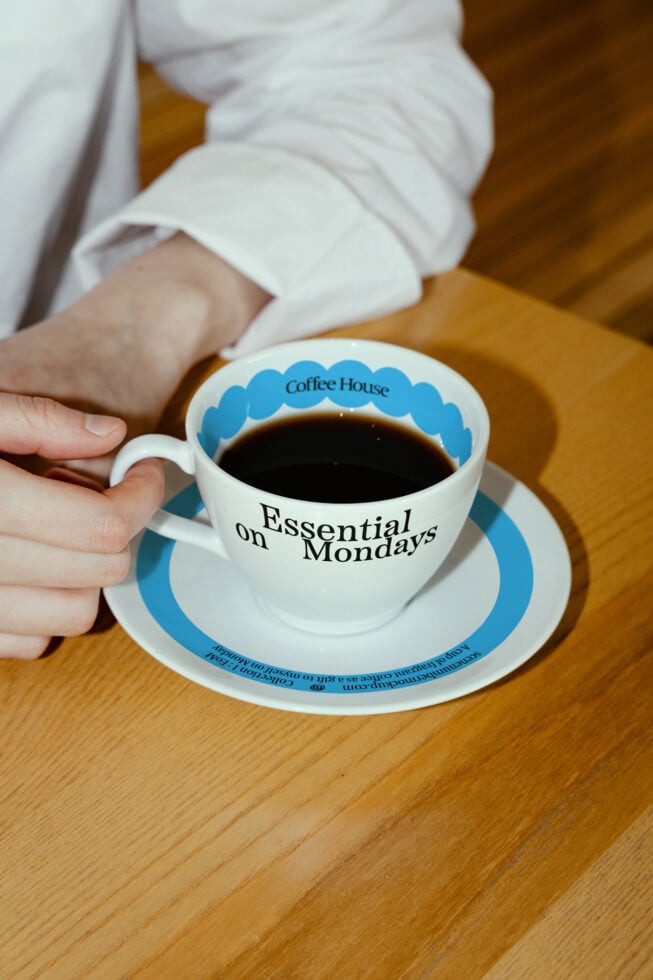 A hand holding a coffee cup on a saucer with Essential on Mondays text. Ideal for mockups templates and graphics. Keywords: coffee cup design mockup.