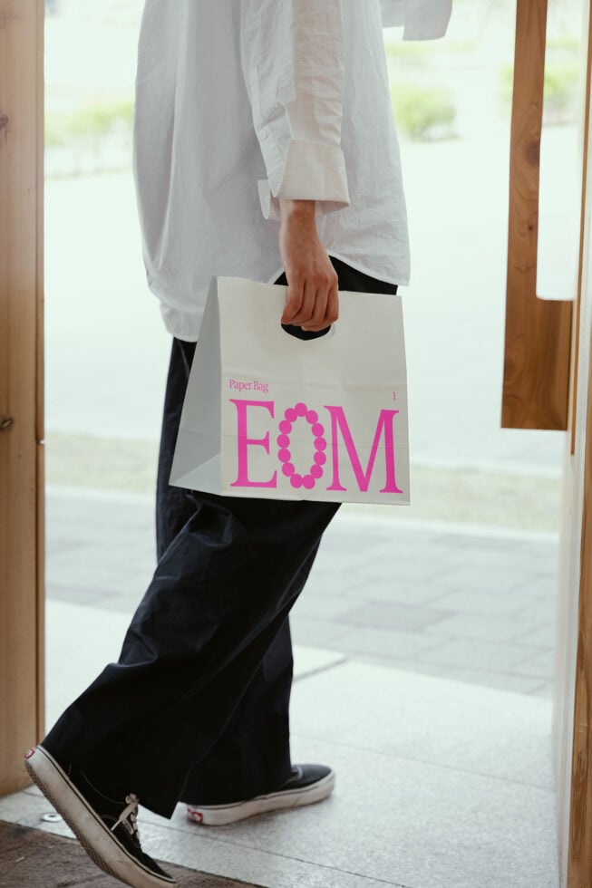 Person holding a white paper bag with pink text EOM walking indoors; stylish packaging mockup; perfect for digital design assets and templates.