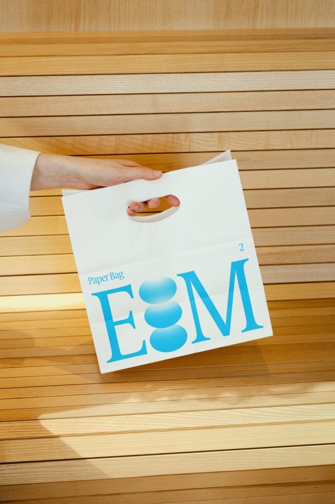 Minimalist paper bag mockup held by hand, featuring modern blue typography on a wooden background. Perfect for showcasing branding designs.