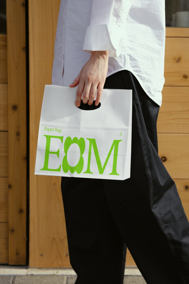 Person holding a white paper bag with green text design. Ideal for product mockup, packaging template, branding, ecommerce, creatives, print design.