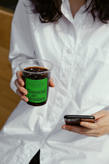 Woman in white shirt holding iced coffee cup with neon green Essential on Mondays label