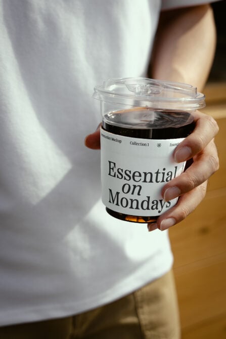 Hand holding clear coffee cup with white label reading Essential on Mondays