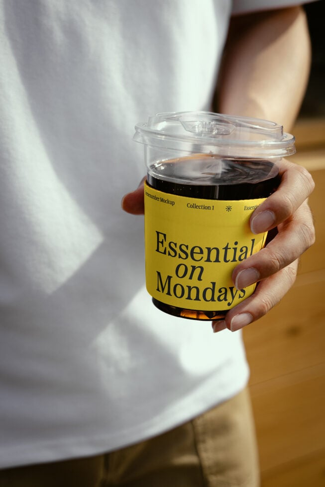 Mockup of a hand holding a coffee cup with a yellow label reading Essential on Mondays. Ideal for beverage branding, packaging design, and marketing templates.