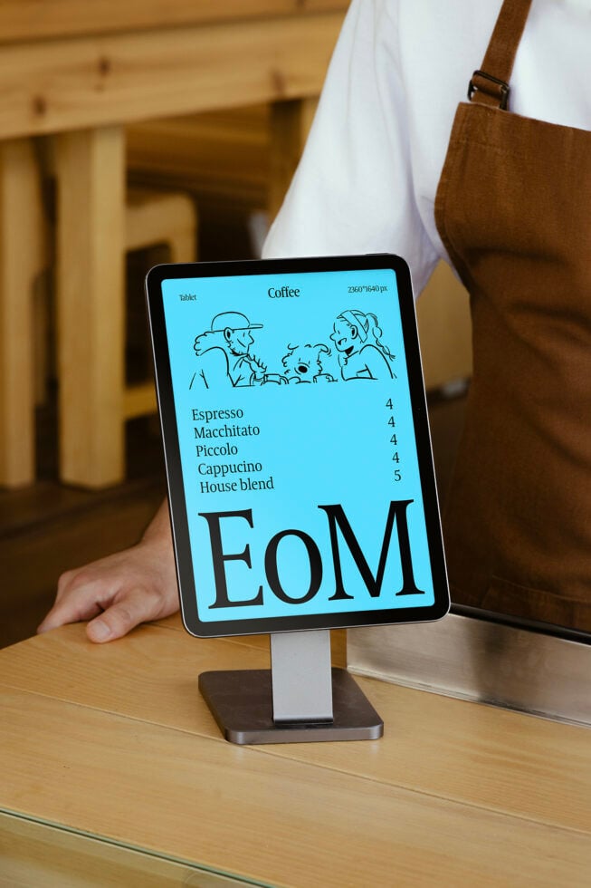 Tablet menu mockup on a wooden counter showcasing a coffee menu with illustration. Keywords: tablet mockup, digital menu, coffee shop design.