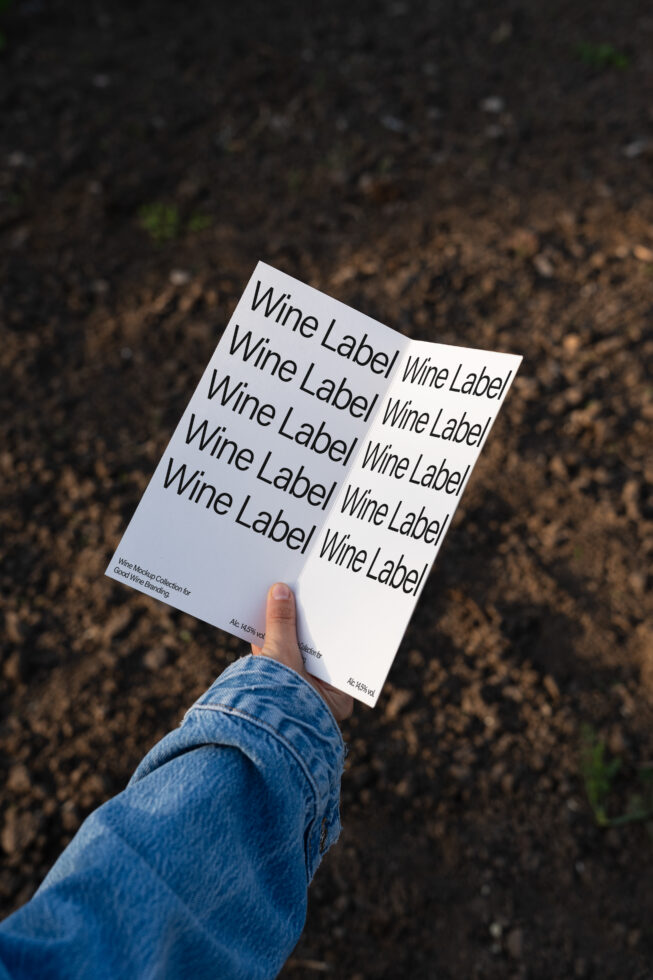 Hand holding a flyer with wine label text design on brown soil background. Ideal for showcasing typography mockups and branding templates for designers.