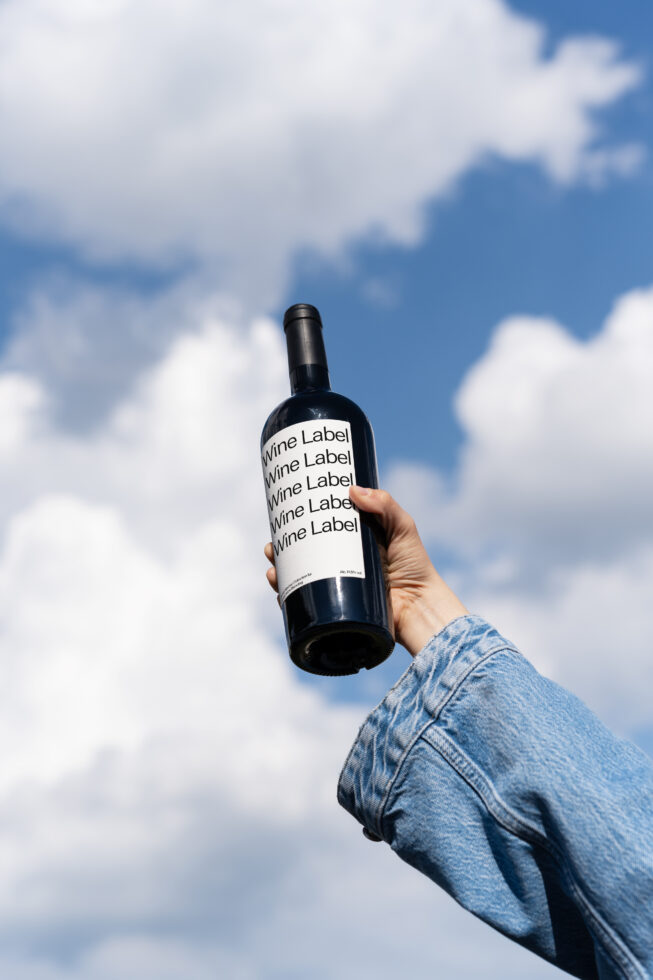 Wine bottle label mockup held by hand in blue denim jacket against blue sky with clouds. Perfect for showcasing branding and design presentations.
