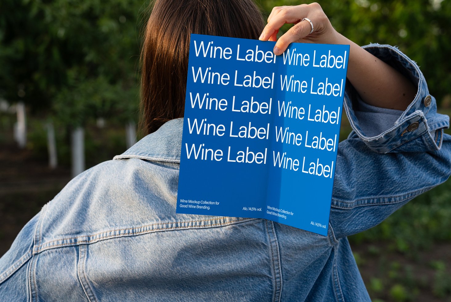 Person holds a blue card featuring repetitive text Wine Label for branding. Ideal for designers seeking wine mockup templates. Denim jacket outdoors.