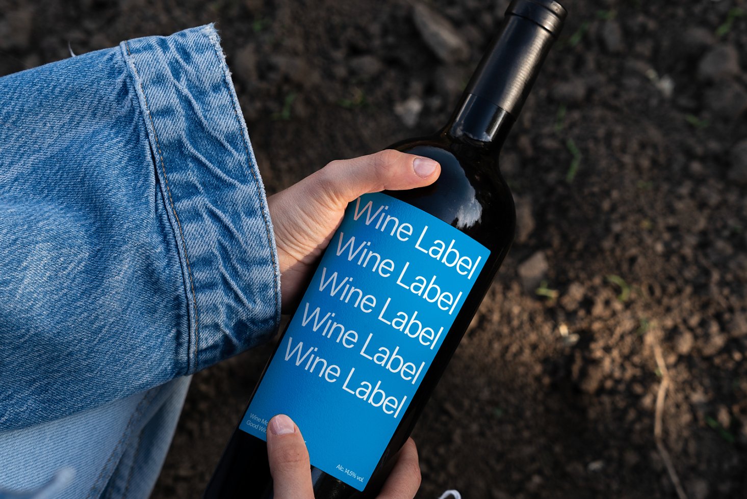 Person holding wine bottle with blue label mockup featuring repeated text Wine Label against outdoor soil background creative design resource.