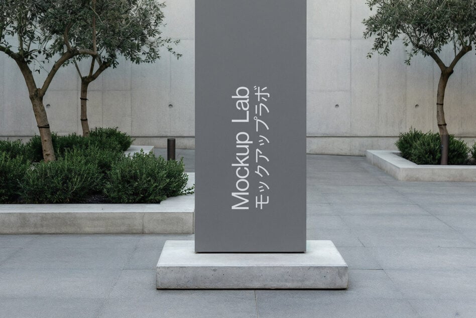 Dark gray vertical sign with white text in courtyard with olive trees