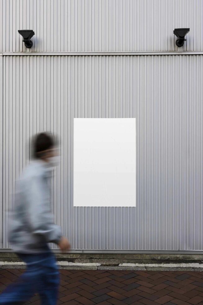 Mockup with a blank poster on a ribbed metal wall, ideal for urban advertising design; a blurred person walks by; suitable for graphic templates.