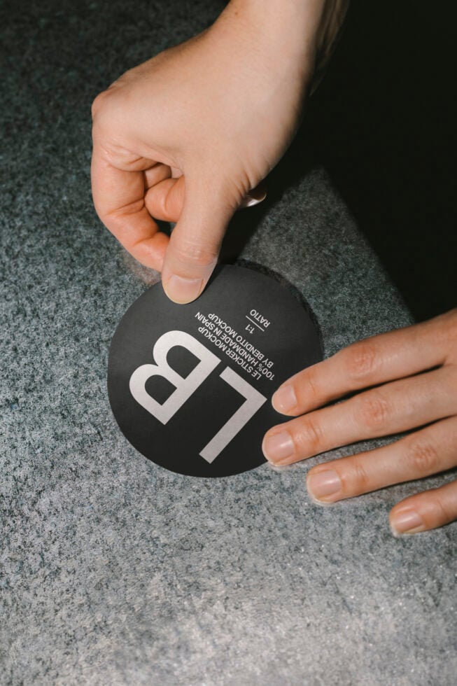 A hand places a round black sticker with bold white text 'LB' on a textured surface. Ideal for branding mockups and design presentations.