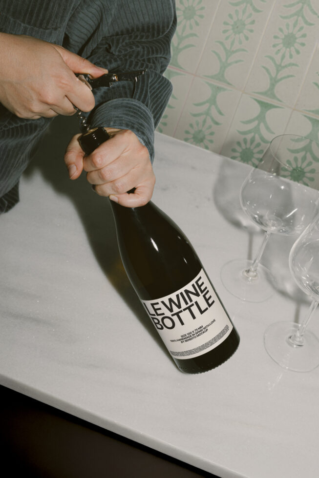 A person opens a wine bottle on a marble countertop with glasses nearby. Perfect for product mockup showcasing in templates or graphics.