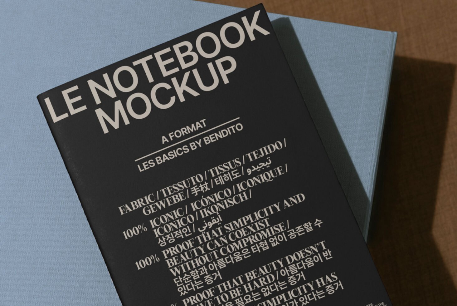 Mockup of a black notebook titled Le Notebook Mockup with multilingual typography placed on a blue surface. Ideal for designers seeking high-quality graphics.