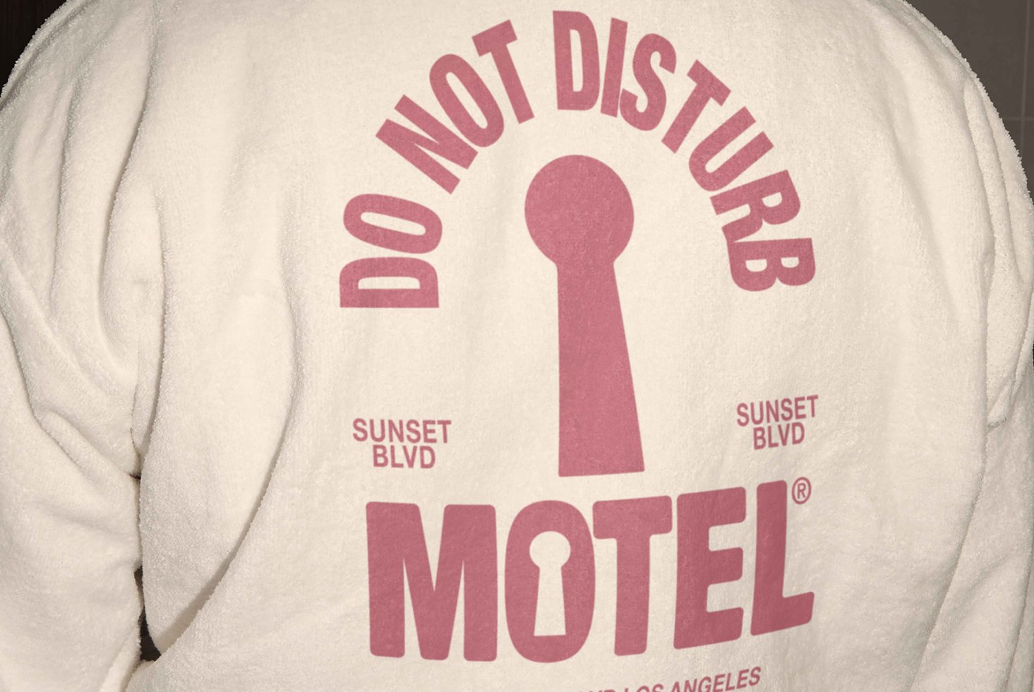 Back of a white robe with pink typography reading Do Not Disturb Motel Sunset Blvd featuring a keyhole graphic. Ideal for fashion design inspiration.