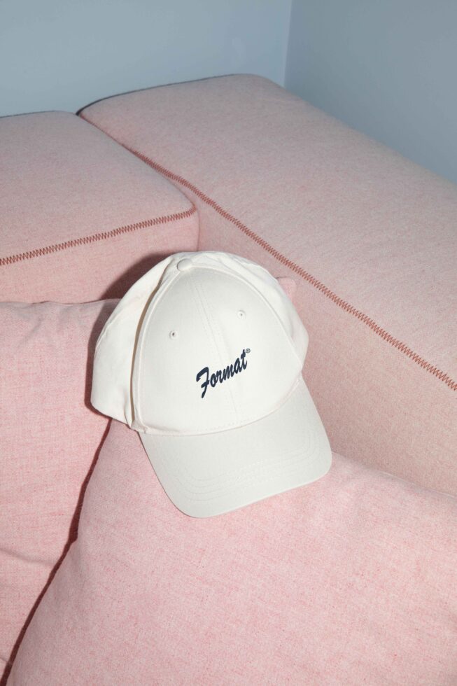 Beige baseball cap with blue text resting on a pink sofa. Minimalist style. Perfect for mockup use in fashion design and branding projects.