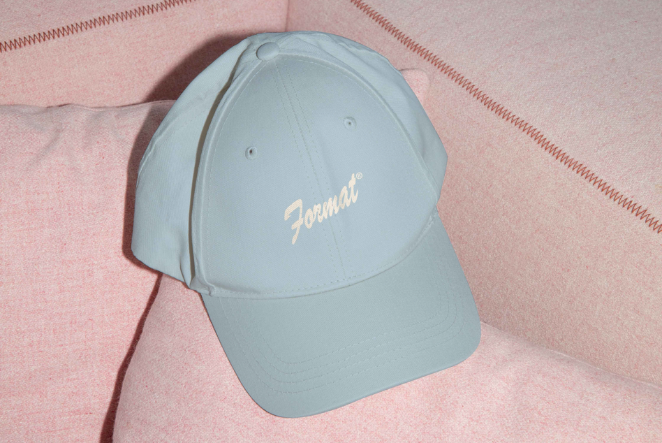 Light blue baseball cap with cream cursive logo on pink fabric background