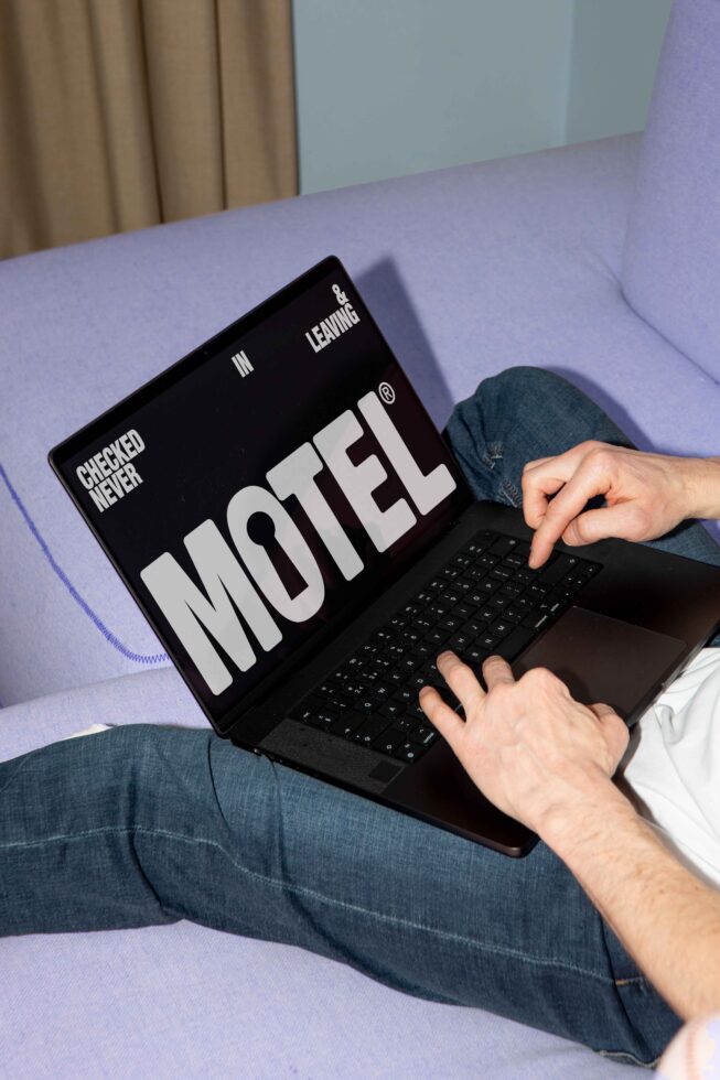 Person on sofa using laptop displaying bold MOTEL text. Ideal for mockups, graphic design resources. Keywords: laptop, typography, digital assets.