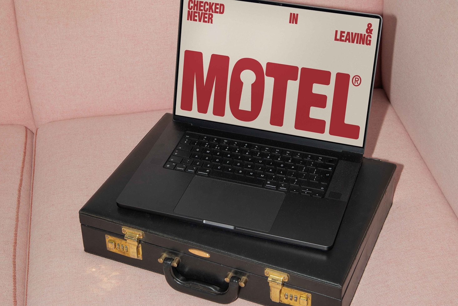 Laptop on a pink couch displaying bold red motel typography on the screen placed on a black briefcase. Ideal for mockups and graphic design projects.