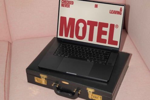 Laptop on a pink couch displaying bold red motel typography on the screen placed on a black briefcase. Ideal for mockups and graphic design projects.