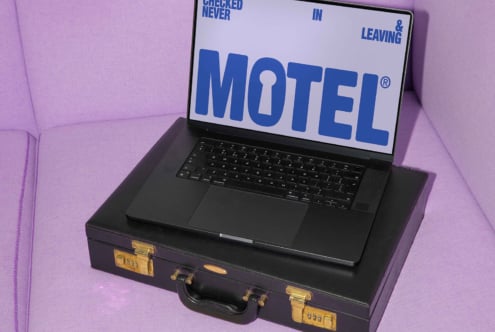 Laptop on suitcase with MOTEL graphic on screen, placed on a purple sofa. Ideal for mockup templates, graphic design assets, digital creatives.
