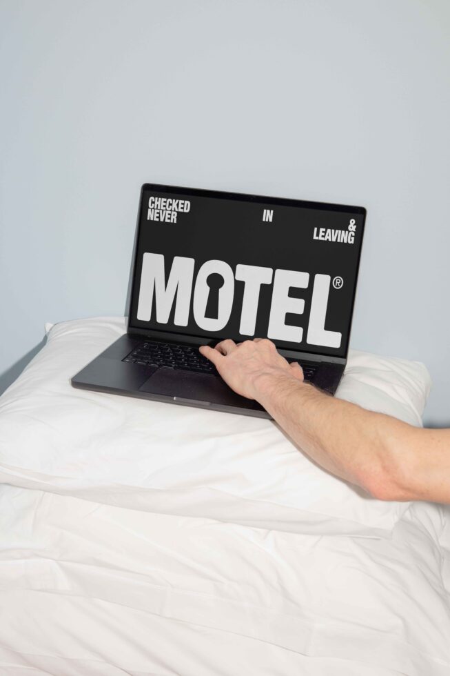 Laptop with bold MOTEL graphic, hand touching keyboard, placed on pillows, suitable for digital assets, mockups, creative templates, graphic design.