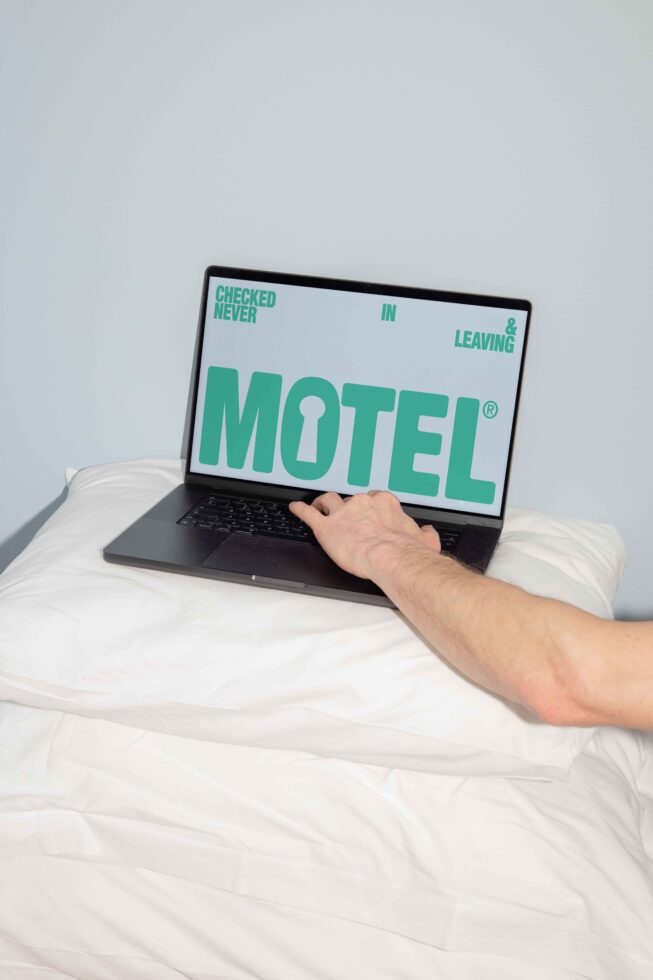 Laptop on a white pillow displaying bold graphic text Motel Checked in never leaving Hand reaching for keyboard digital design mockup concept deco