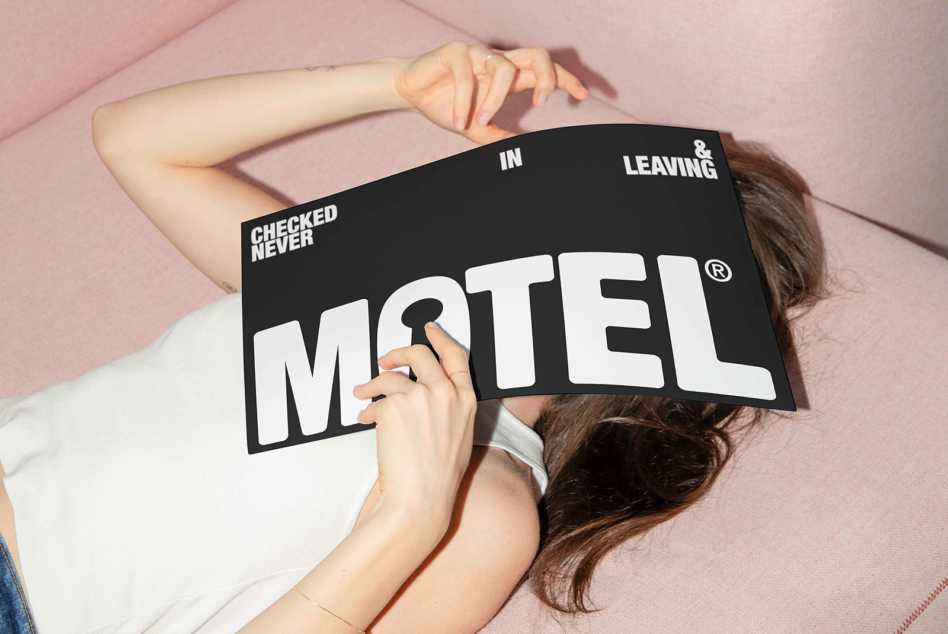 Person lying on pink couch holding motel sign reading checked never in leaving