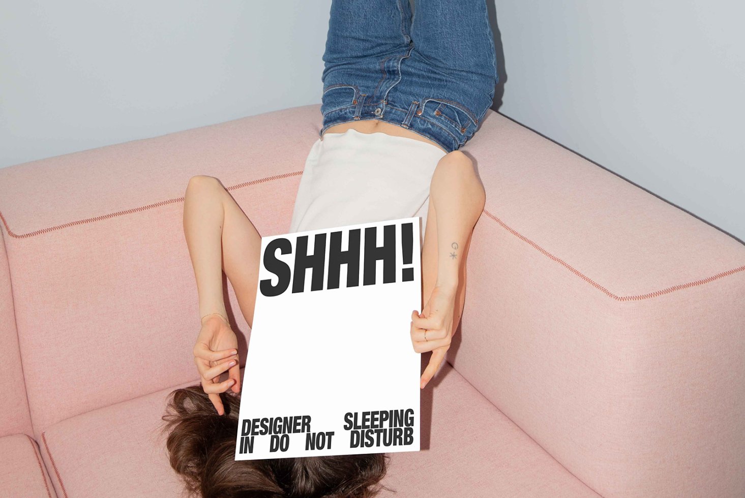 Designer laying on pink couch holding SHHH sign for mockup. Casual jeans outfit. Ideal for creative studio marketing. Typography and decor concept.
