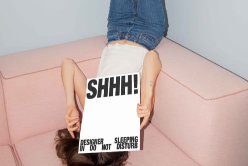 Designer laying on pink couch holding SHHH sign for mockup. Casual jeans outfit. Ideal for creative studio marketing. Typography and decor concept.