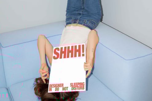 Person lying on blue sofa upside down holding sign that says Shhh Designer Sleeping Do Not Disturb modern mockup photography creative design resource