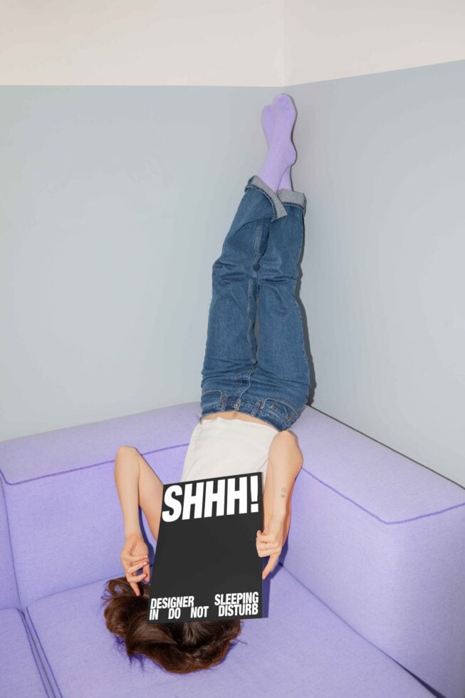 Designer lying upside down on a purple couch with a large black SHHH sign covering face. Perfect for graphic design templates and creative mockups.