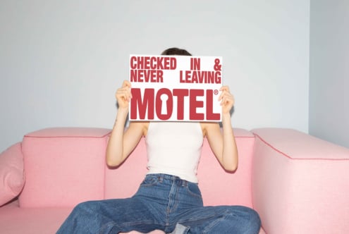Woman holding motel sign on pink sofa beside light gray wall. Ideal for graphics designers seeking creative inspiration in casual home settings.