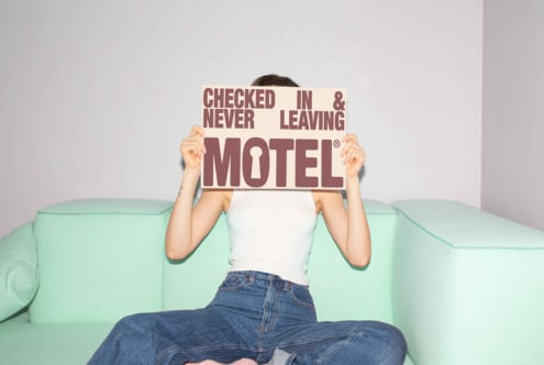 Person holding sign Checked In Never Leaving Motel sitting on mint green sofa. Casual outfit. Poster mockup for designers interior graphic template.