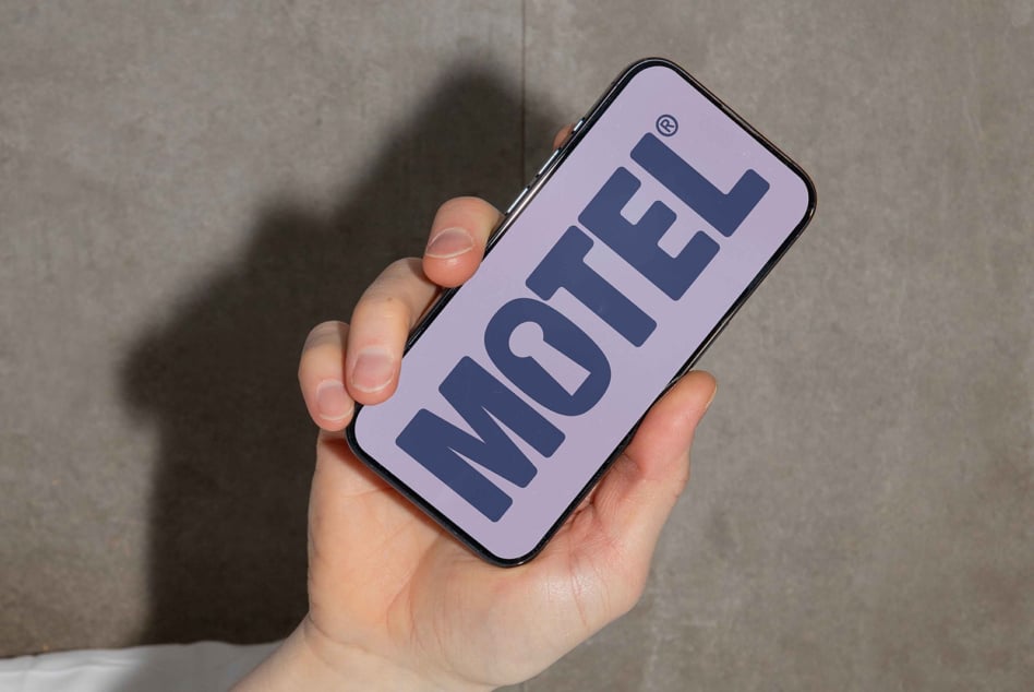 Close-up of a hand holding a smartphone displaying a bold sans-serif font spelling MOTEL on a lavender screen. Perfect for font design mockups.