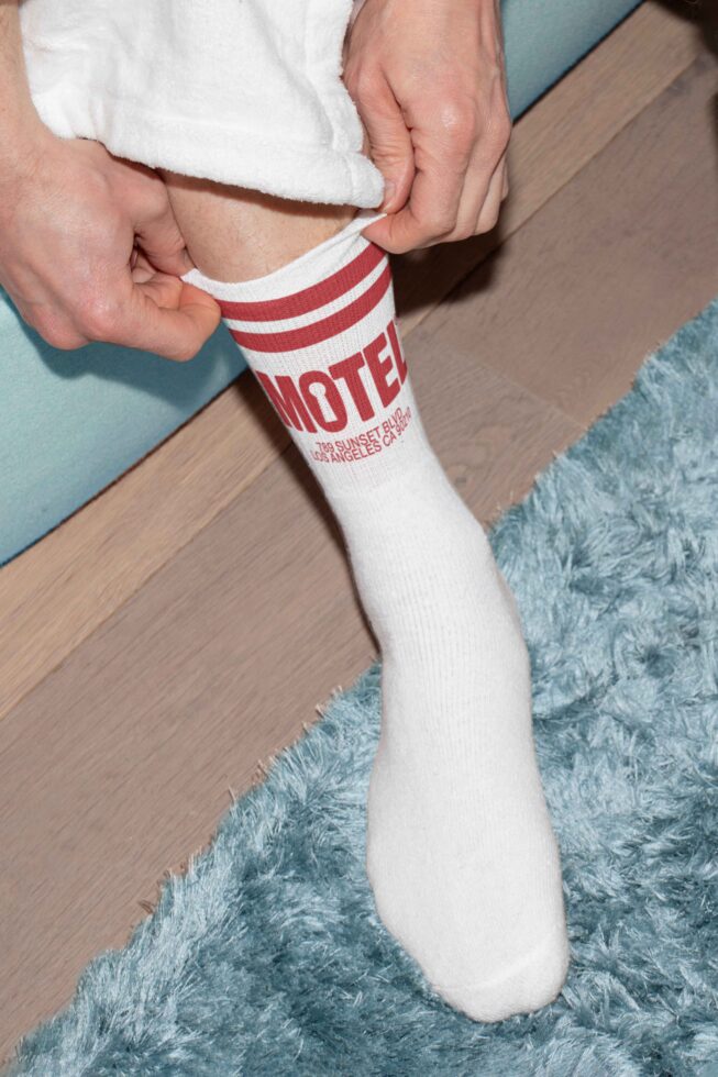 Close-up of hands adjusting white socks with red motel logo and text Los Angeles on a wooden floor with blue rug. Ideal for mockup or graphic use.
