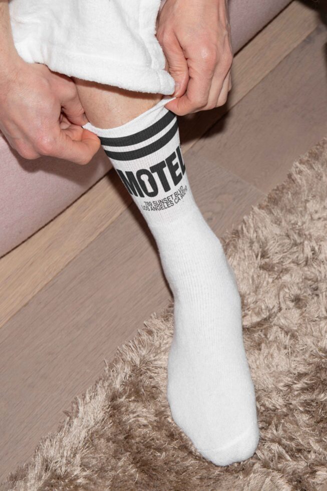Person adjusting white sock with black stripes and Motel design logo while sitting on a beige carpet. Suitable for mockups and graphic design projects.