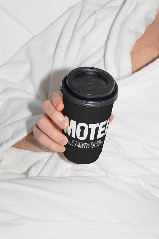 Person holding a black coffee cup with branding lying on a white bed, design mockup, coffee branding, packaging, lifestyle, designers resource