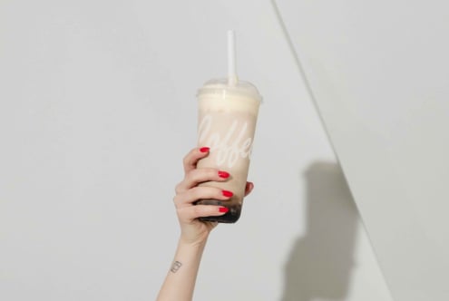 Hand holding a bubble tea cup with red nails and tattoo visible against a neutral background ideal for mockup designs and beverage branding usage.