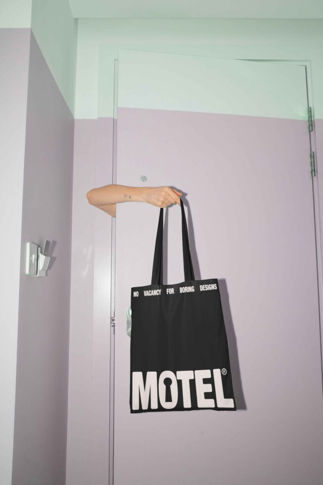 Black tote bag with bold text Motel held by a hand against a pastel purple door background perfect for design mockups and branding presentations.