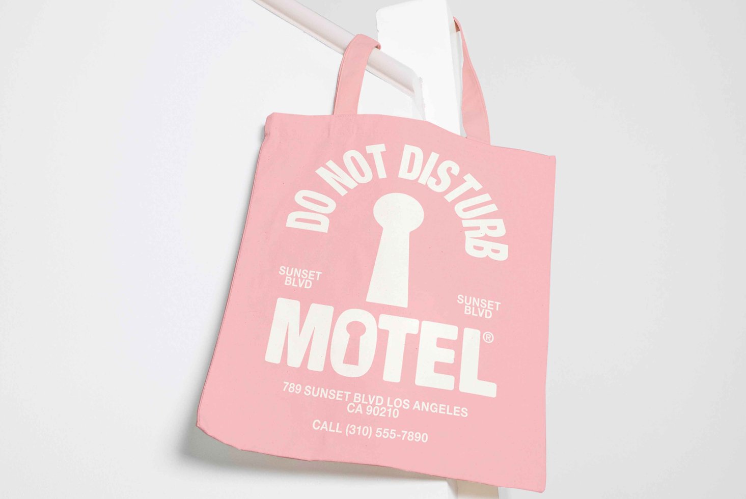 Pink tote bag mockup with white DO NOT DISTURB text and motel details hanging on a white surface. Perfect for graphic designers and branding templates.