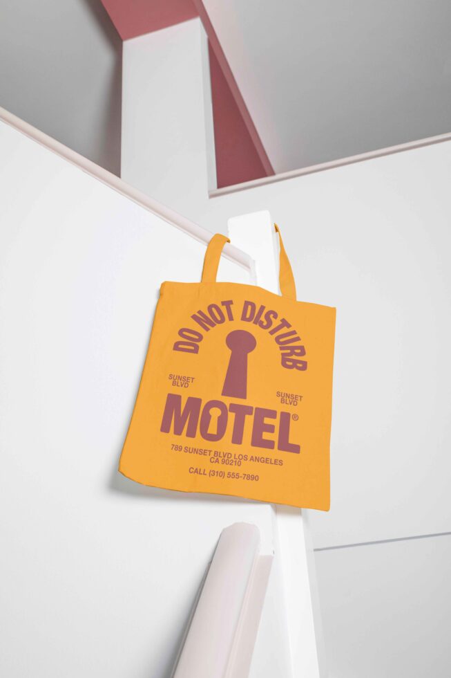 Yellow tote bag with vintage motel graphics hanging on pink and white wall, ideal for mockup design, travel-themed graphics, retro vibe templates.