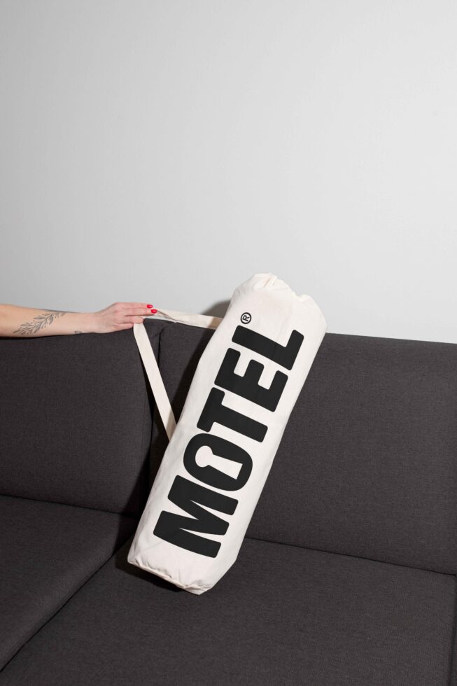 White tote bag with bold black font text Motel on a gray sofa not showing a person's face perfect for mockup and branding design project display.