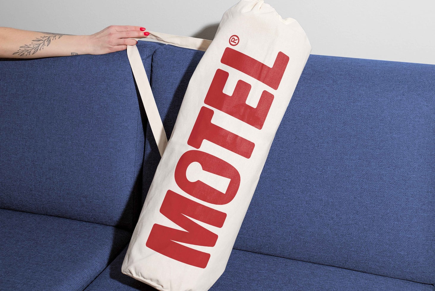 Canvas tote bag with bold red Motel font design held by hand on a blue sofa; perfect for typography mockups, branding graphics, and designer templates.